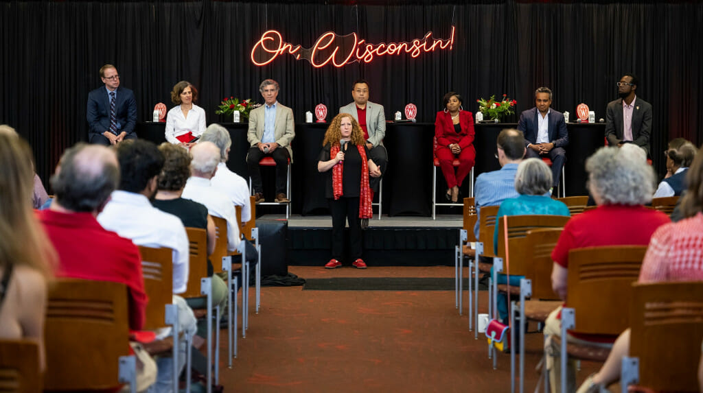 Chancellor Mnookin talks to a crowd while standing in front of a panel of UW–Madison faculty and alumni seated on a stage.