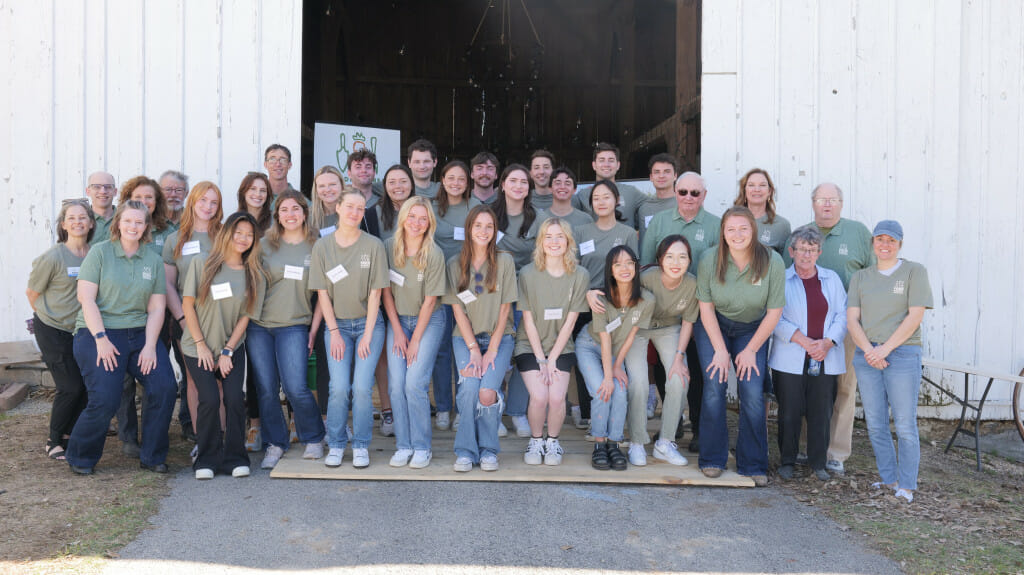 A group of college students and staff from Madison Area Food Pantry Gardens stand in three rows outside of a farm outbuilding. They are wearing matching green T-shirts and jeans.
