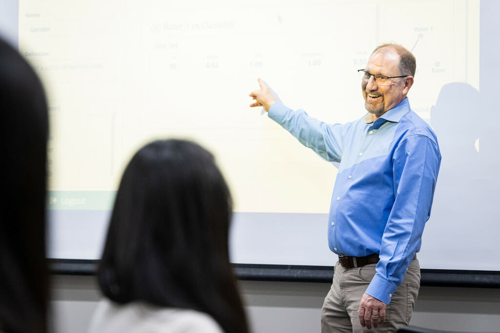 A man gestures to a whiteboard as students listen.