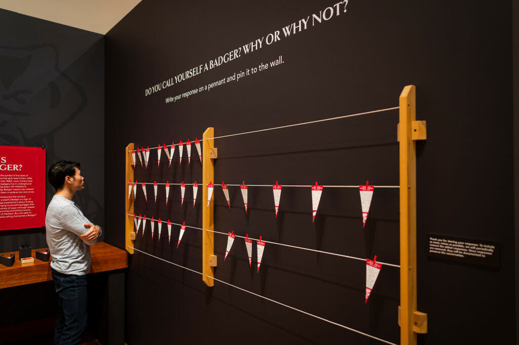 A man looks at a wall display in a gallery space. On the wall, paper pennants are pinned to string. Above the display, words on the wall read, "Do you call yourself a Badger? Why or why not? Write your response on a pennant and pin it to the wall."