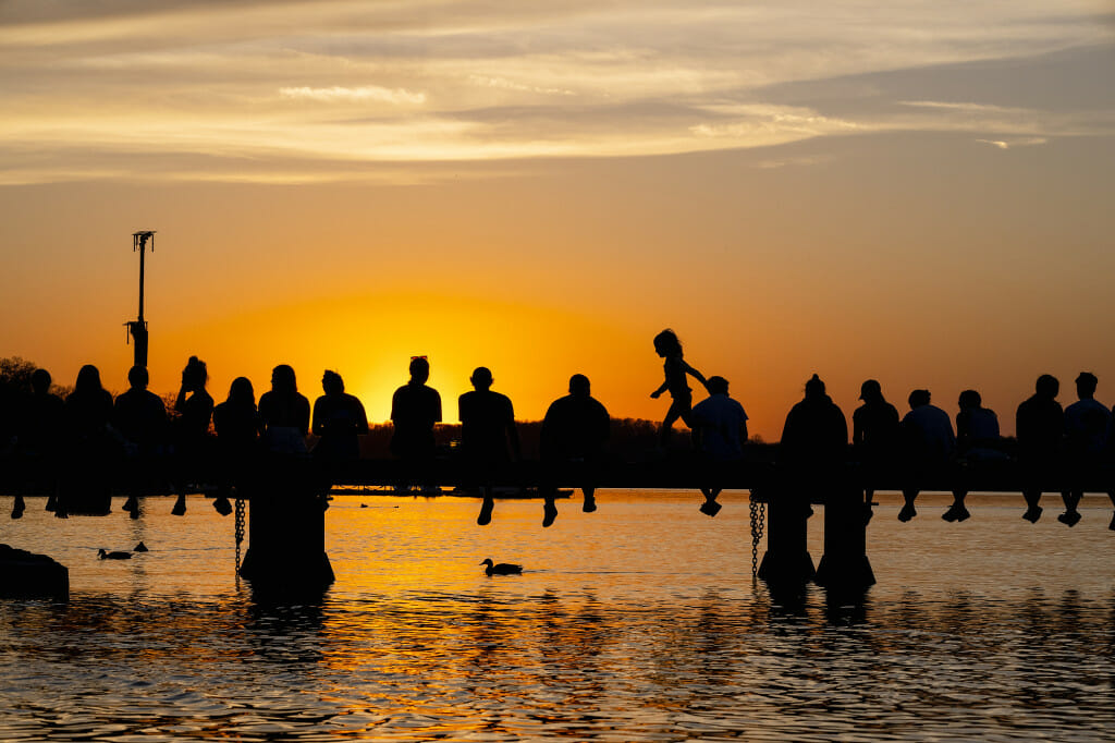 People sitting on a pier over a lake are silhoutted by a sunset.