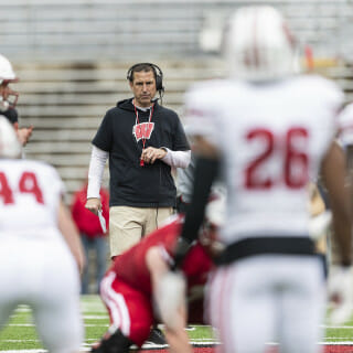 The camera focuses on Coach Fickell in a black polo shirt, with white-clad football players in the foreground.