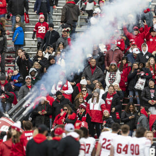 Smoke shoots out of a t-shirt gun as fans stand in the stands and hold their arms up.