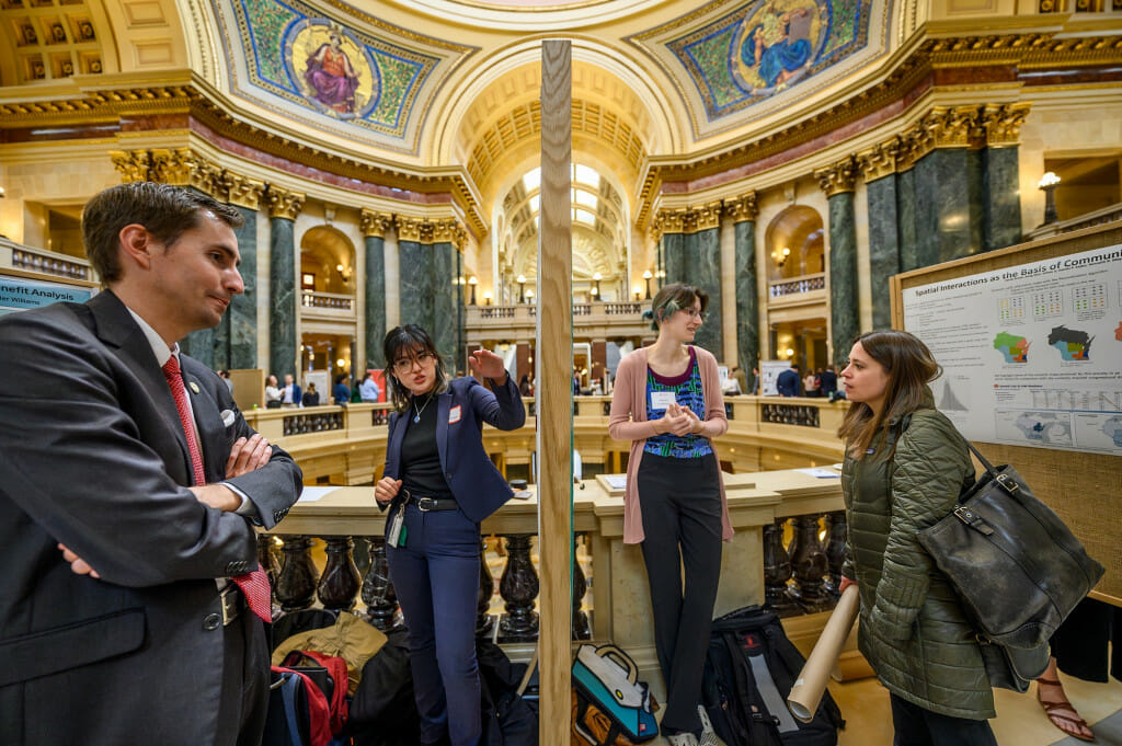 A large poster display splits the shot of two people explaining their research posters in the Wisconsin Capitol rotunda.