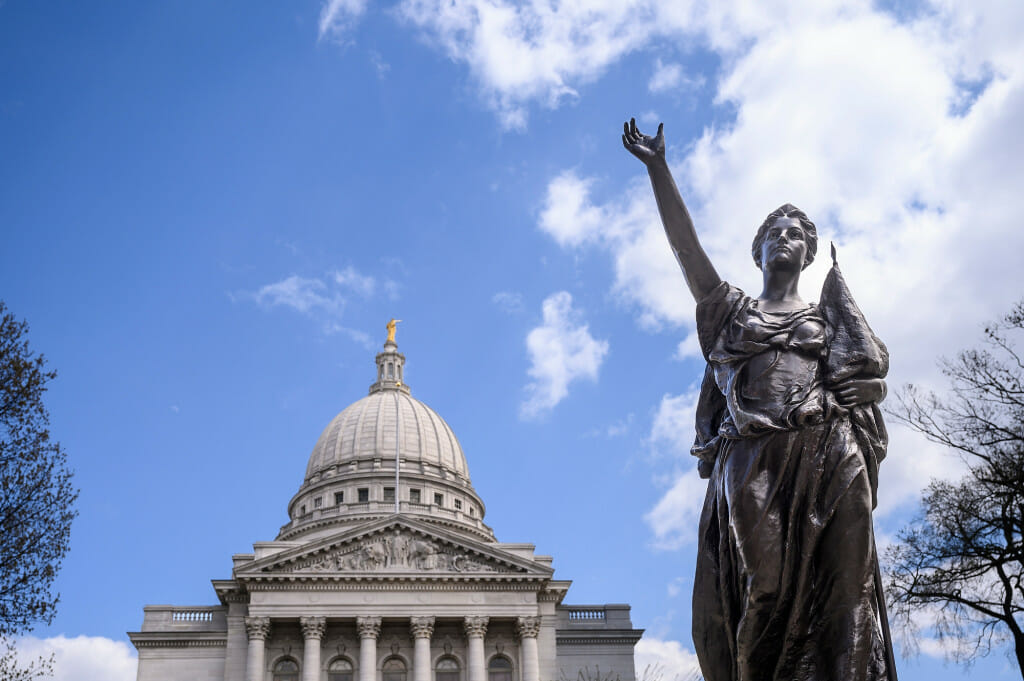 The bronze statue of a woman representing Wisconsin's state motto “Forward” reaches toward the sky while the Wisconsin State Capitol can be seen in the background