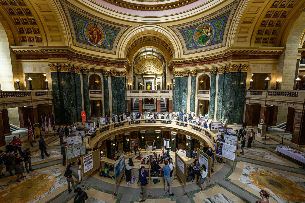 A wide view of the Wisconsin Capitol rotunda, a neoclassical space in green and white marble, filled with people attending a research poster session.