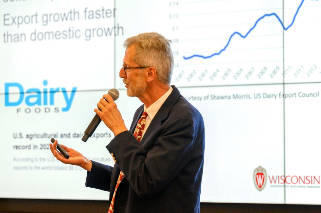 A man holds a microphone and gestures with his free hand as he explains the information seen on a projection screen behind him.