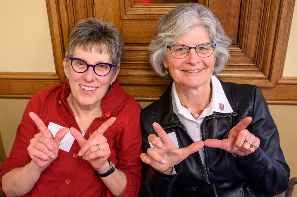Two women raise their hands to make a W sign with their thumbs and forefingers. They are smiling to the camera.