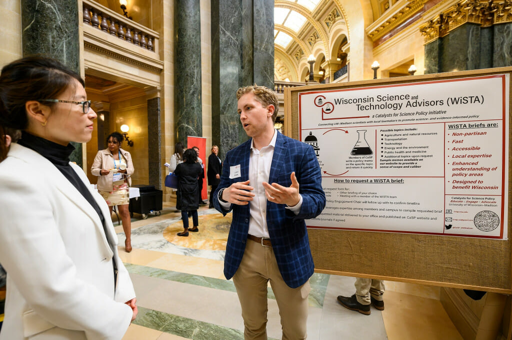 A man gestures with his hands as he explains the content of a research poster on display in the Wisconsin Capitol rotunda.