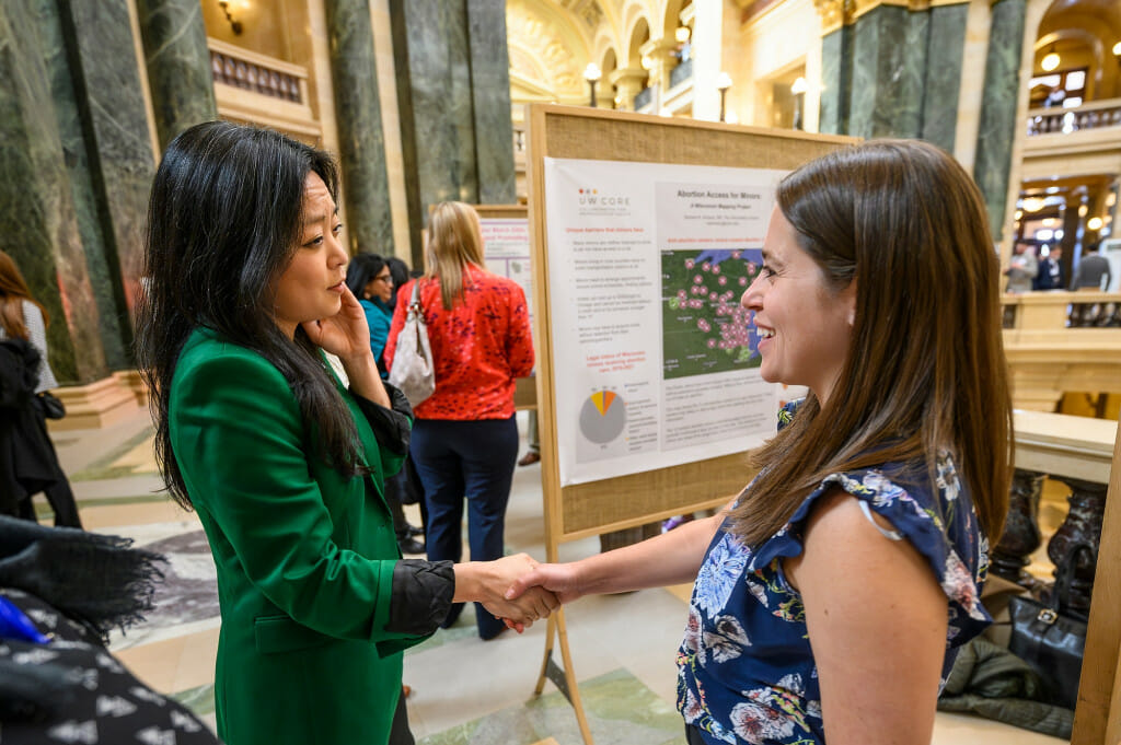 A woman in a green suit coat shakes hands and smiles with a second woman in the Wisconsin Capitol rotunda.