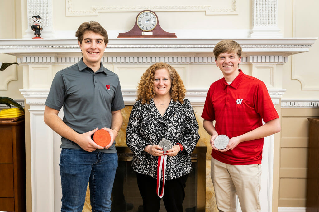 Mechanical Engineering undergraduate students Teekay Kowalewski (left), Jennifer Mnookin (center) and Dylan Zinkgraf (right) face the camera, standing in front of a white, neoclassical fireplace with a clock on the mantle. Kowalewski holds a 3D-printed version of the medallion. Mnookin holds the new, pewter medallion. Zinkgraf holds an aluminum rendition of the medallion.