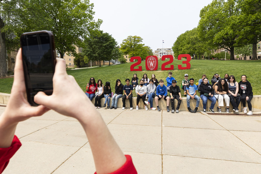 Students stand in front of a red 2023 sign on a green lawn leading up to a majestic building.