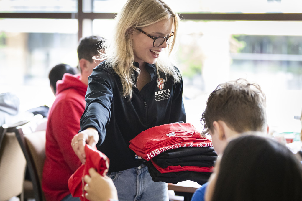 A smiling woman picks up a t-shirt from a pile and hands it to a student.