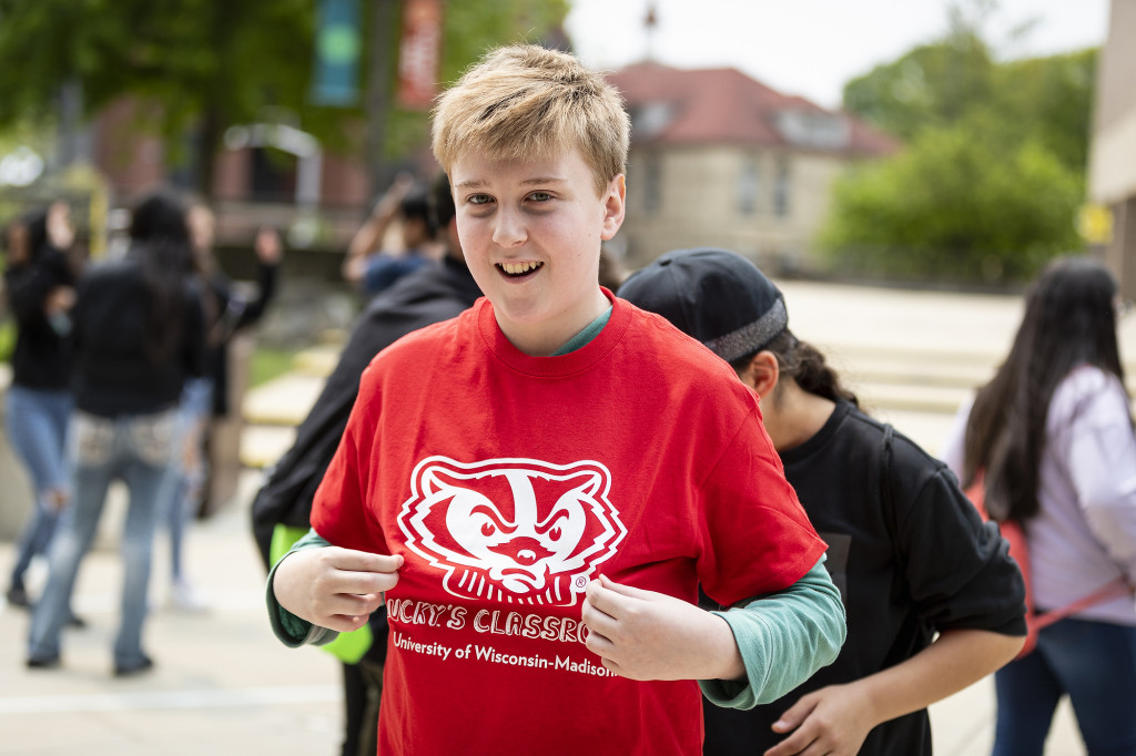 A boy in a red t-shirt depicting Bucky Badger smiles and holds out tteh front of it to display it.