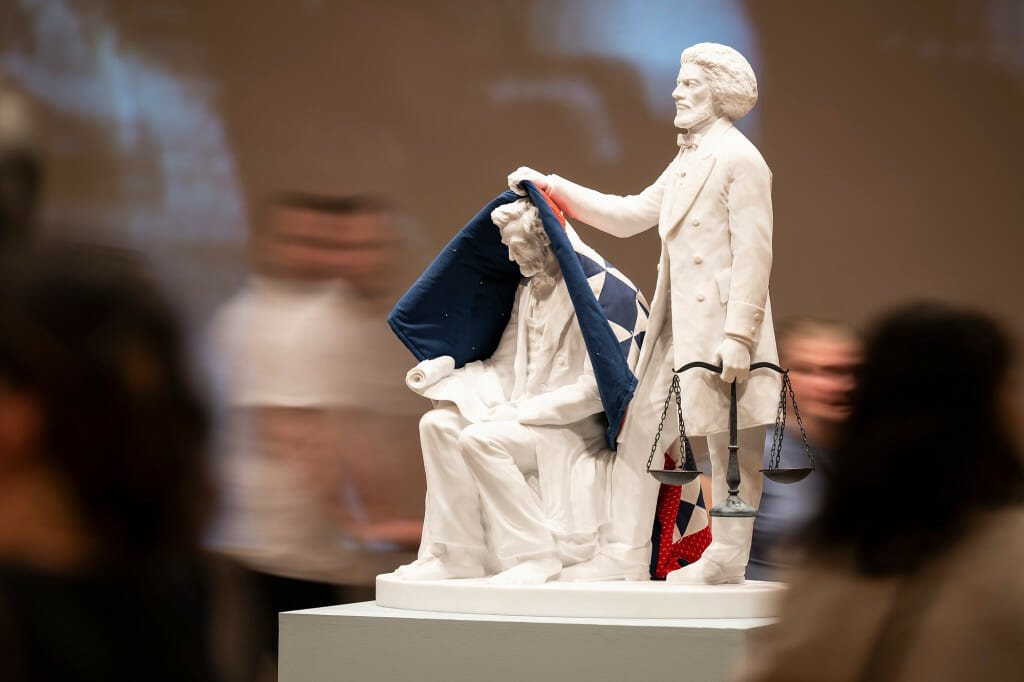 A statue depicts one man removing a blanket off another man.