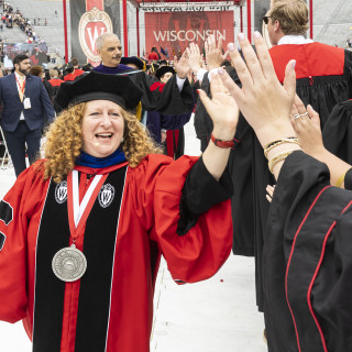 A woman in red academic regalia high fives several graduates wearing commencement robes.