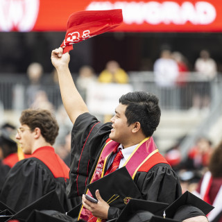 A man in a commencement robe waves a red tool as others around him cheer.