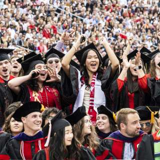 People in graduation robes raise their hands and celebrate, in the bleachers at Camp Randall.