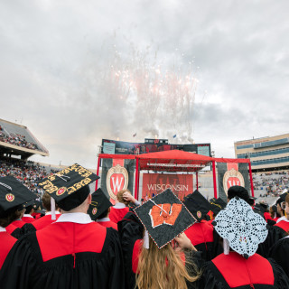 The back of five graduates with decorated mortarboards is seen in the foreground, with fireworks in the background.