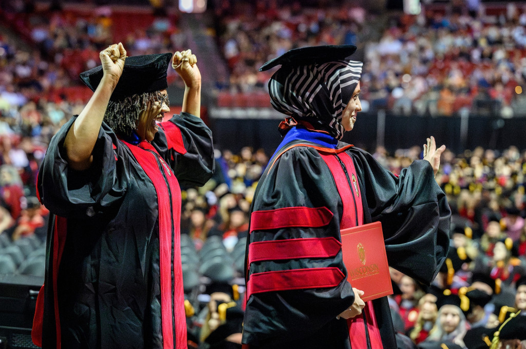 Two people in commencement robes cross the stage, one cheers the other.