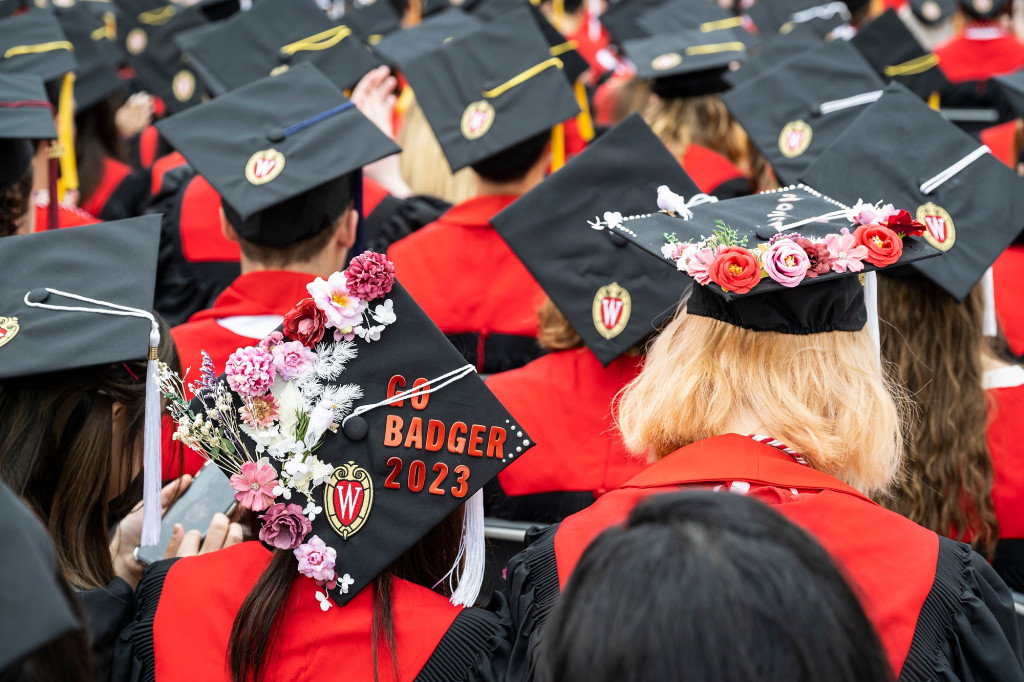 Two women wear mortarboard caps decorated with flowers, one reads 