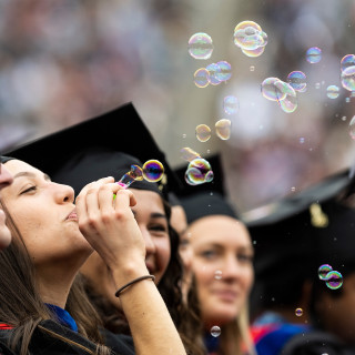 A woman blows bubbles as people in commencement robes are all around her.