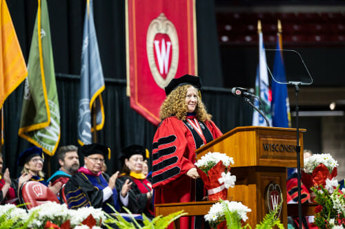 Wearing cap and gown, Jennifer Mnookin stands at a podium and speaks to an unseen audience. Behind her, academic dignitaries applaud.