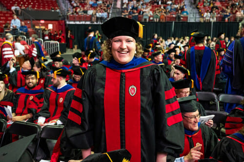 Graduate Hanna Barton smiles at the camera. They are wearing academic regalia and standing amid a crowded arena of fellow graduates in caps and gowns.