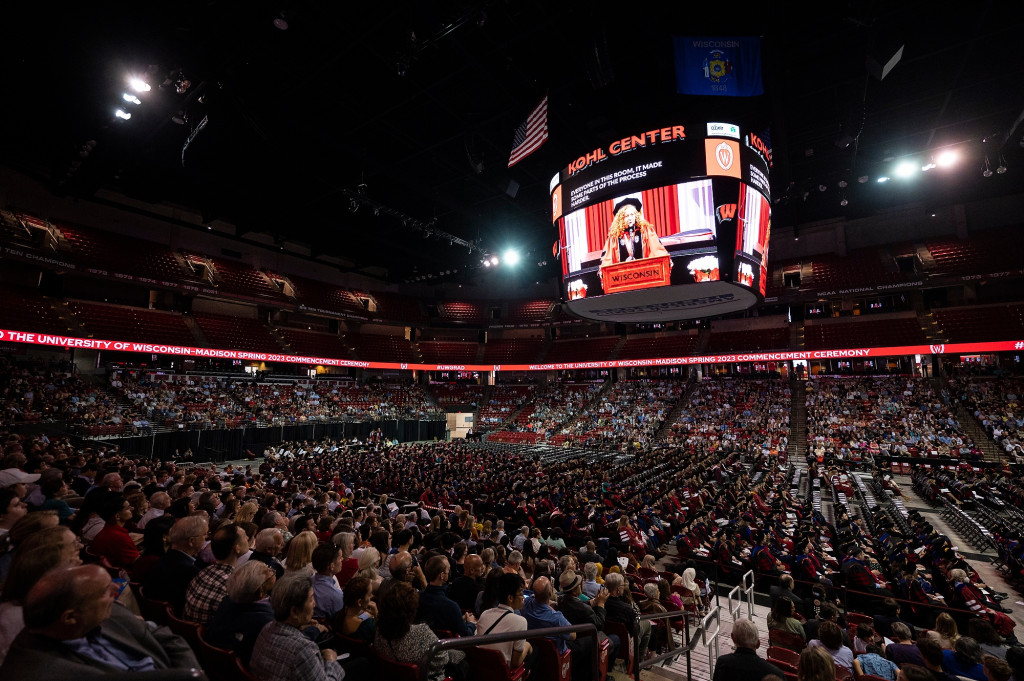 The interior of the Kohl Center, crowded with people and graduates, is shown, with chancellor Jennifer Mnookin projected on the big screen.