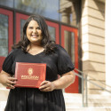 Yasmeena Ougayour stands outside the School of Education, a sandstone building with white columns and red doors. She is smiling to the camera and holding a red diploma folio with the University of Wisconsin–Madison seal and the words 
