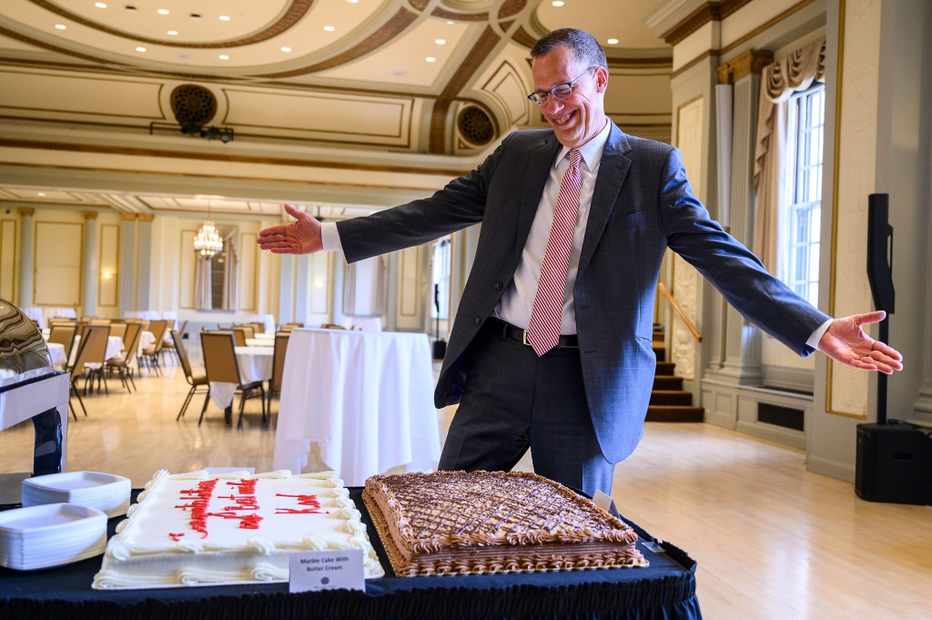 A man in a suit and tie smiles and stretches his arms wide in appreciation of a German chocolate cake.