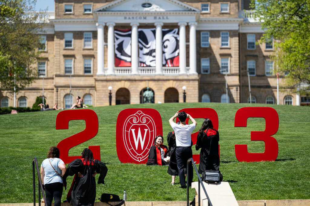 A sweeping view of Bascom Hill with red 2023 numbers at the bottom, with people in graduation gowns posing with them.