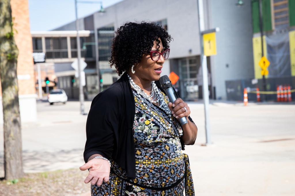 A woman holds a microphone and speaks in the street.