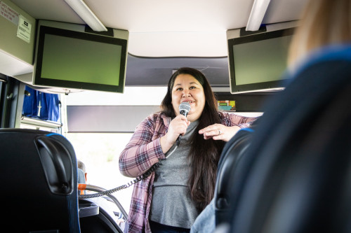 A woman stands at the front of a bus and talks into a microphone.