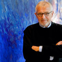 A photo portrait of Francis Halzen standing in front of a blue abstract mural