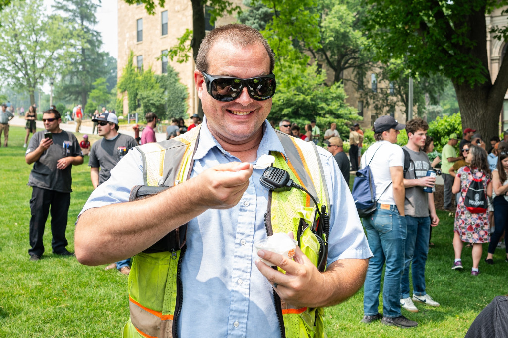 Tom Staskal smiles as he scoops a bite of ice cream. He is wearing sunglasses and a neon safety vest with a radio pinned to his shirt.