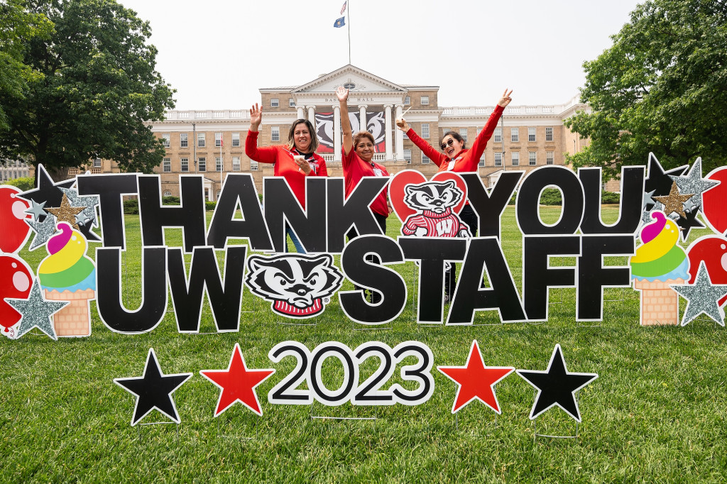 Three women in red shirts raise their hands in celebration and smile at the camera. They're standing on Bascom Hill with Bascom Hall behind them and a big lawn sign in front of them that reads Thank You UW Staff 2023.