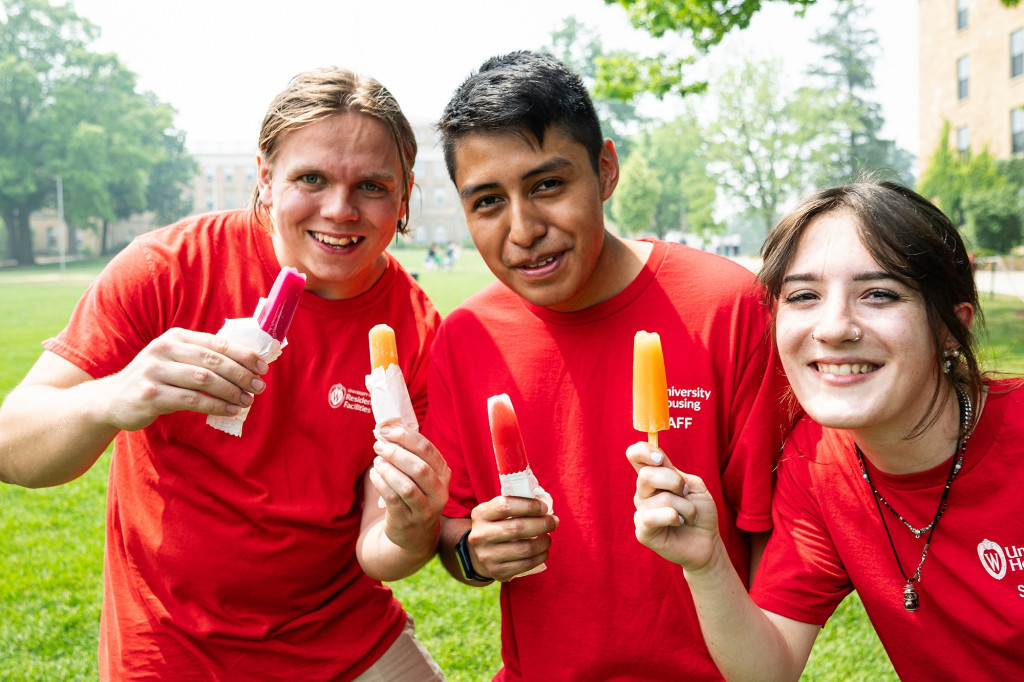 Three undergraduate students wearing red UW Housing Staff T-shirts hold up popsicles and smile during a Staff Appreciation Ice Cream Social on Bascom Hill.