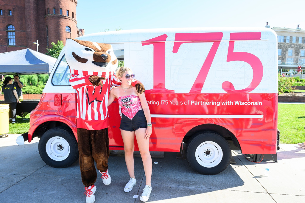 Bucky Badger and a woman wearing a UW tank top pose for a photo outside an ice cream truck painted red and white with the words 175 - Celebrating 175 Years of Partnering with Wisconsin.