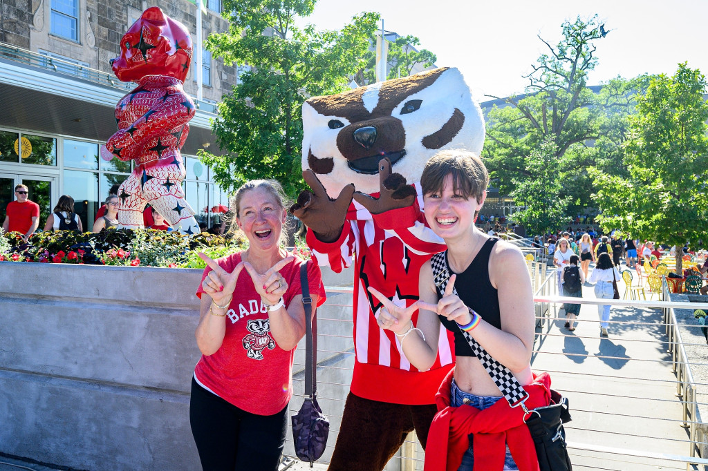 Two people wearing badger red smile and make W signs with Bucky Badger. The new Bucky on Parade statue is behind them.