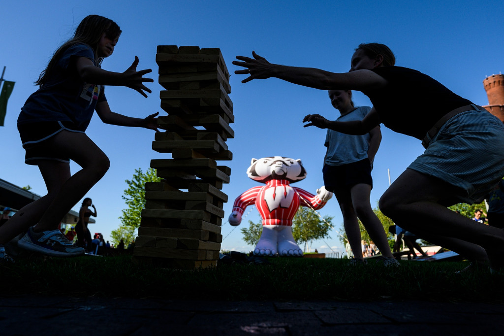 Kids backlit against the blue sky play a giant game of Jenga. A larger-than-life inflatable Bucky Badger looks down on the scene.