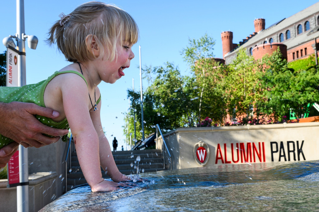 Held up by an adult, a toddler leans into a large fountain at Alumni Park.