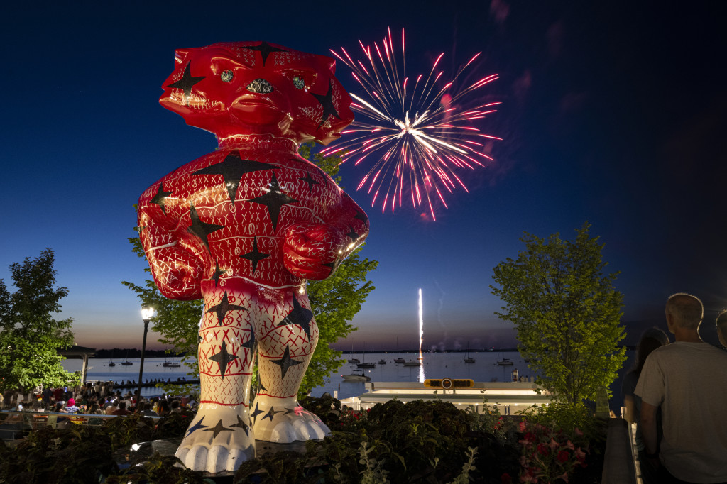 A colorful red, white and blue Bucky Badger statue stands proudly while a single firework explosion bursts behind him over Lake Mendota.