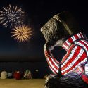 A statue of Bucky Badger is posed sitting with his cheek resting on his hand. He is looking away toward Lake Mendota where a fireworks display lights the sky.