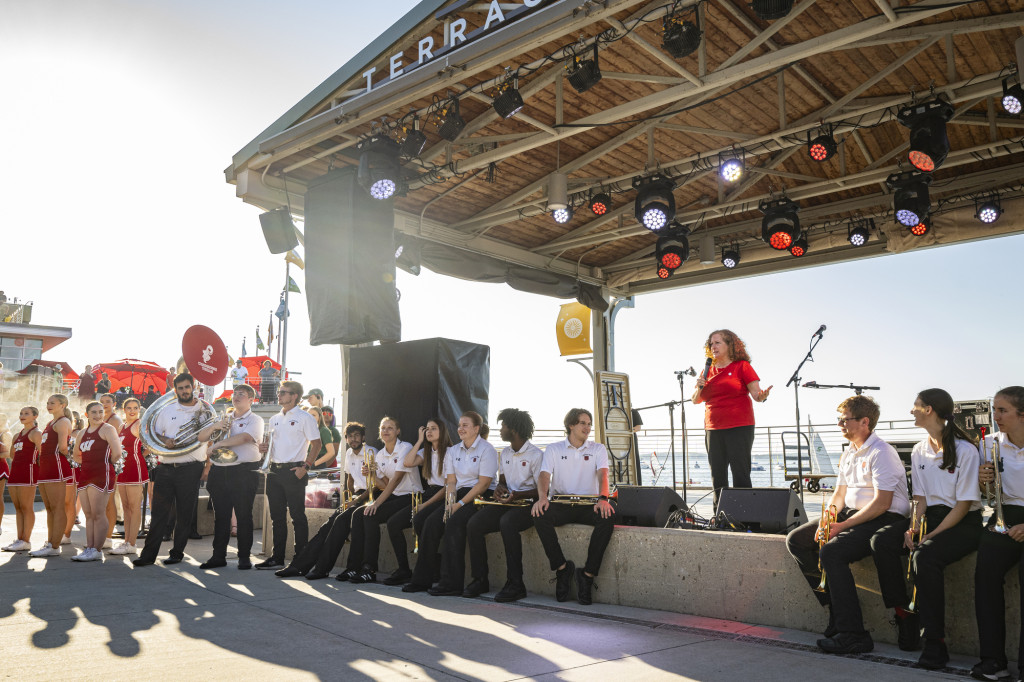 Chancellor Jennifer Mnookin stands on the stage at Memorial Union Terrace and speaks to the crowd. Members of the UW Marching Band are seated on the stage edge in front of her.