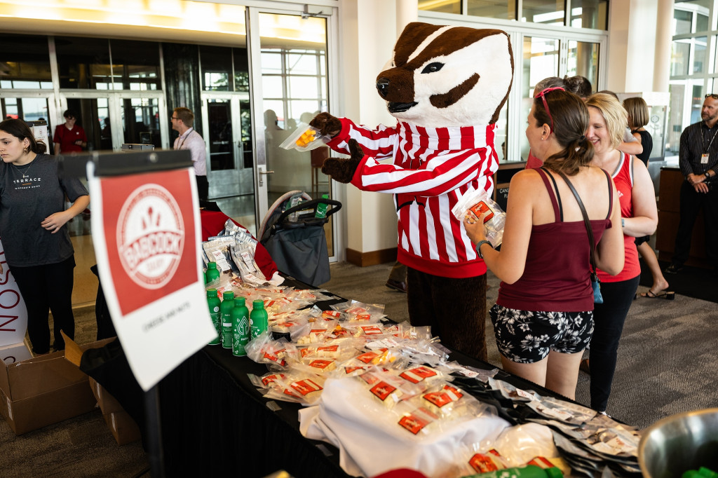 Indoors, Bucky Badger holds up a bag of cheese curds as people take food and drink from a snack table.