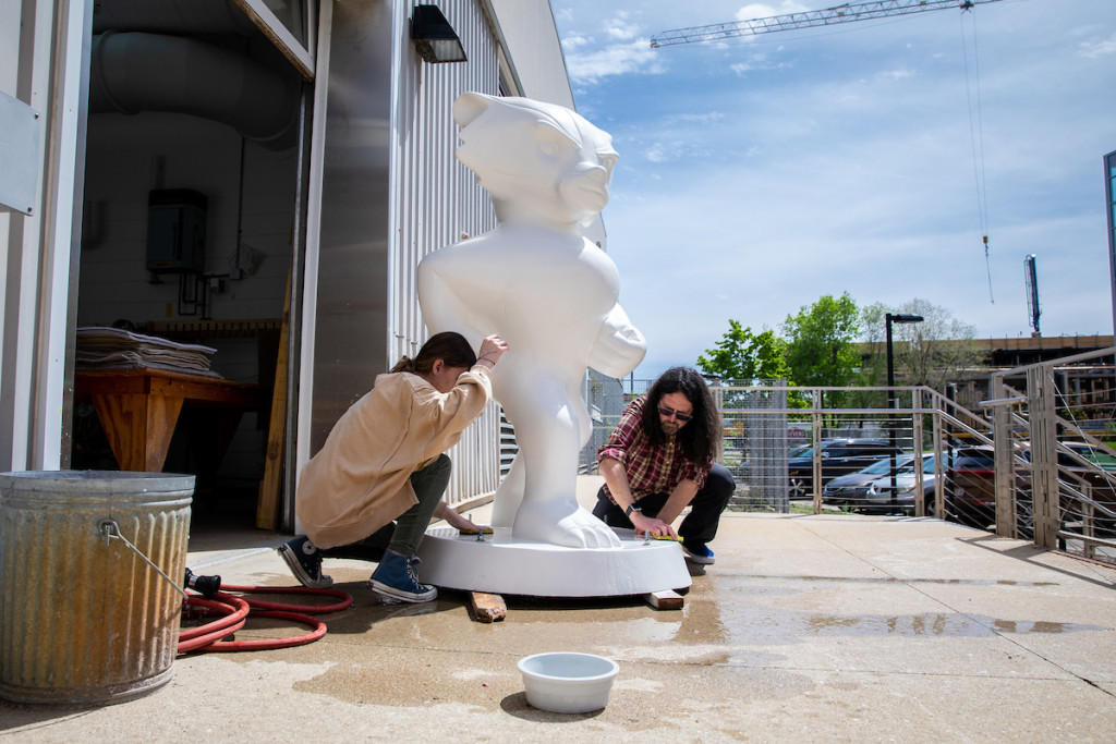 Two people work on a plain white Bucky Badger statue in a parking lot.