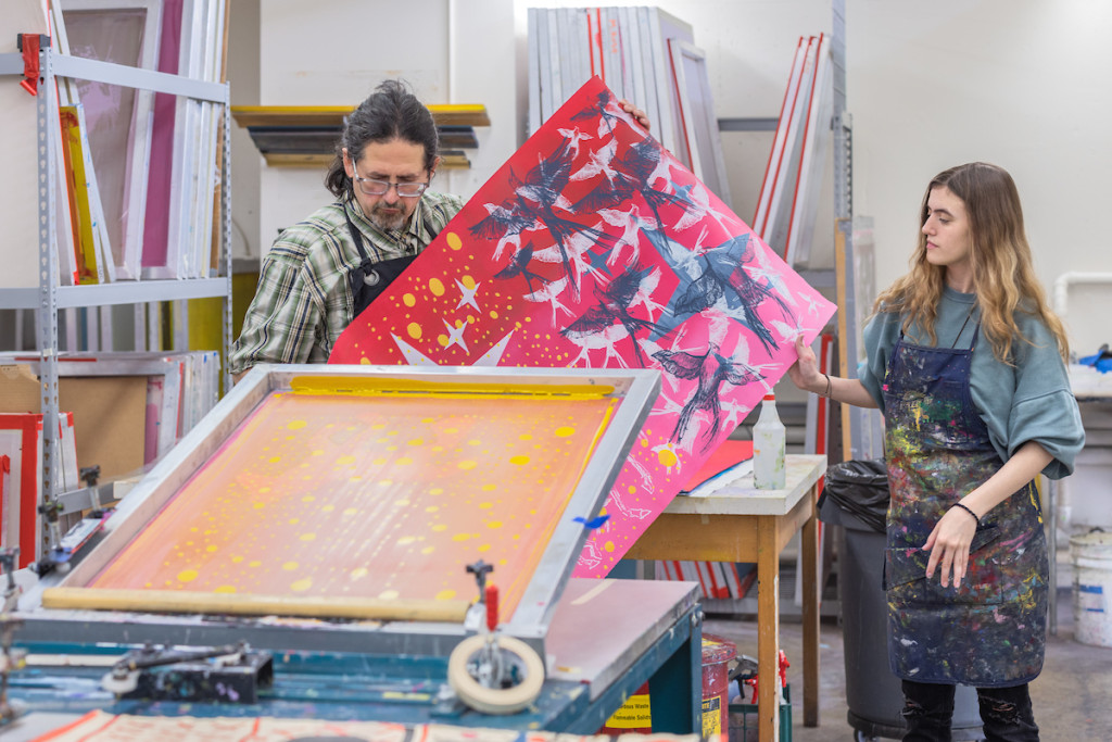 A man withdraws a colorful print from a printmaking machine.