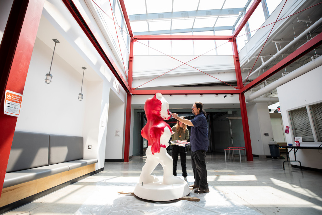 Two people apply red paint to a white Bucky Badger statue in an empty hall.