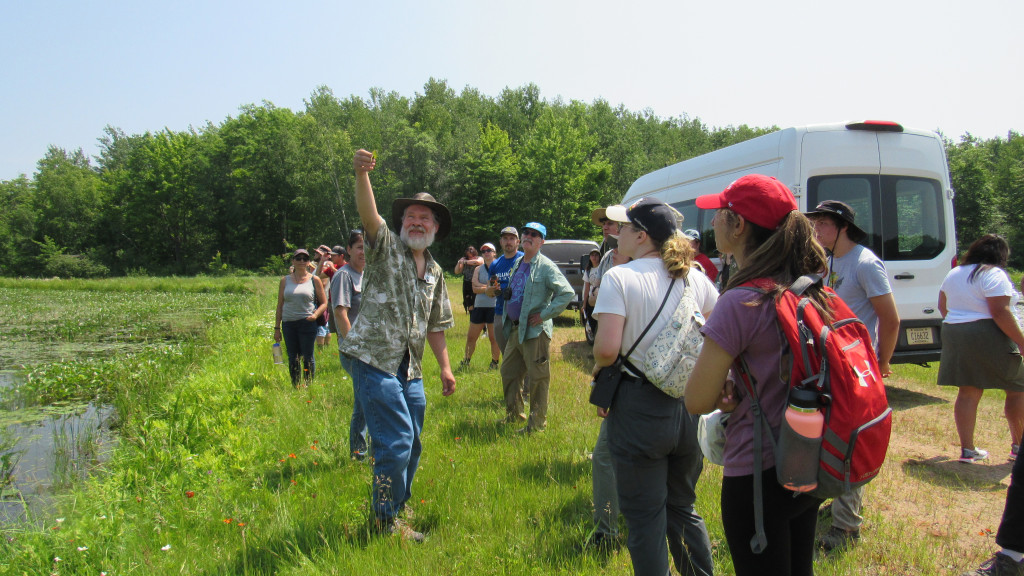 A man with a white beard and brimmed hat holds a plant sprig to the light for a group of college students and instructors. They are standing in short grass next to a wetland.
